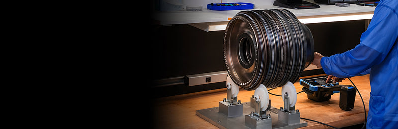 A large metallic turbine component mounted on a testing fixture, with a person in a blue lab coat operating equipment at the right side in a laboratory setting.